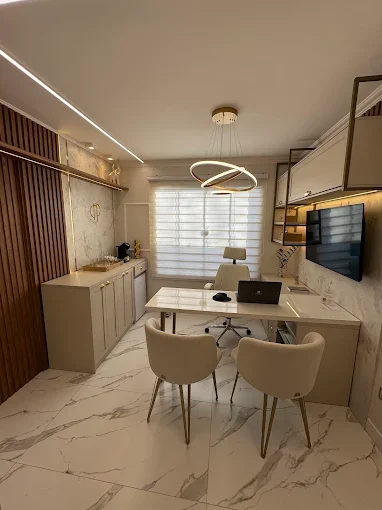 Consultório da Dra. Pamela Paula com mesa branca, poltronas bege, luminária circular dourada e piso de mármore